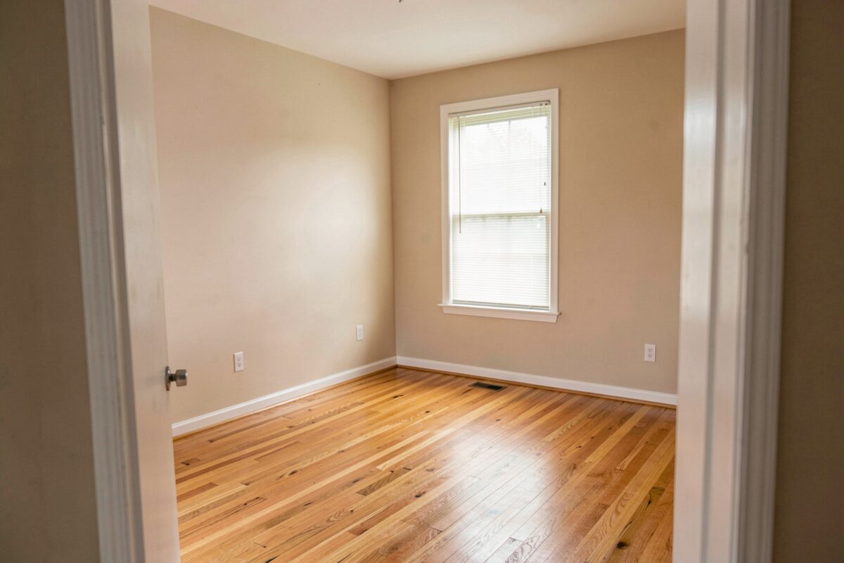 An empty bright rental room photographed from the doorway, bare hardwood floor, a single window with blinds, warm neutral walls, no furniture