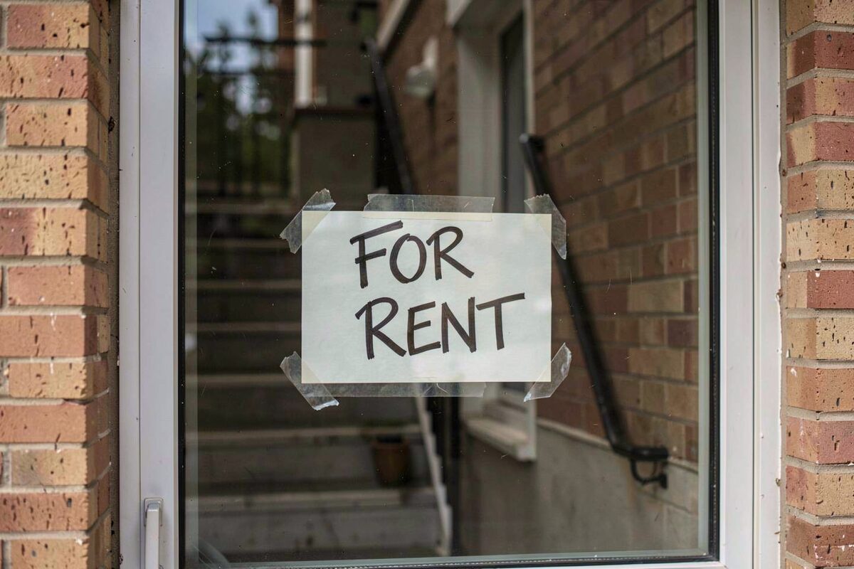A hand-lettered For Rent sign taped inside the front window of a brick low-rise apartment building, the sign slightly askew, a staircase visible dimly behind the glass